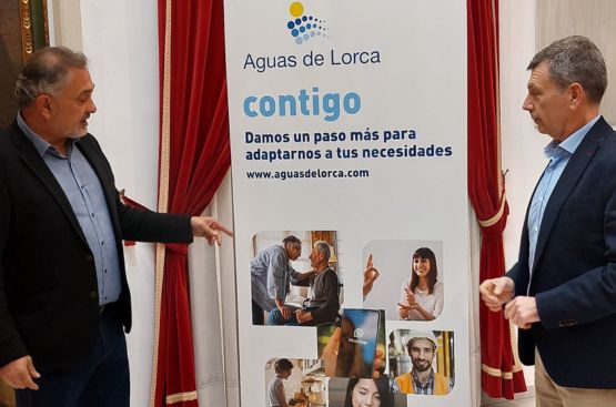 Rueda de prensa con el concejal Ángel Meca y el gerente de Aguas de Lorca señalando un cartel con la información del Programa Contigo